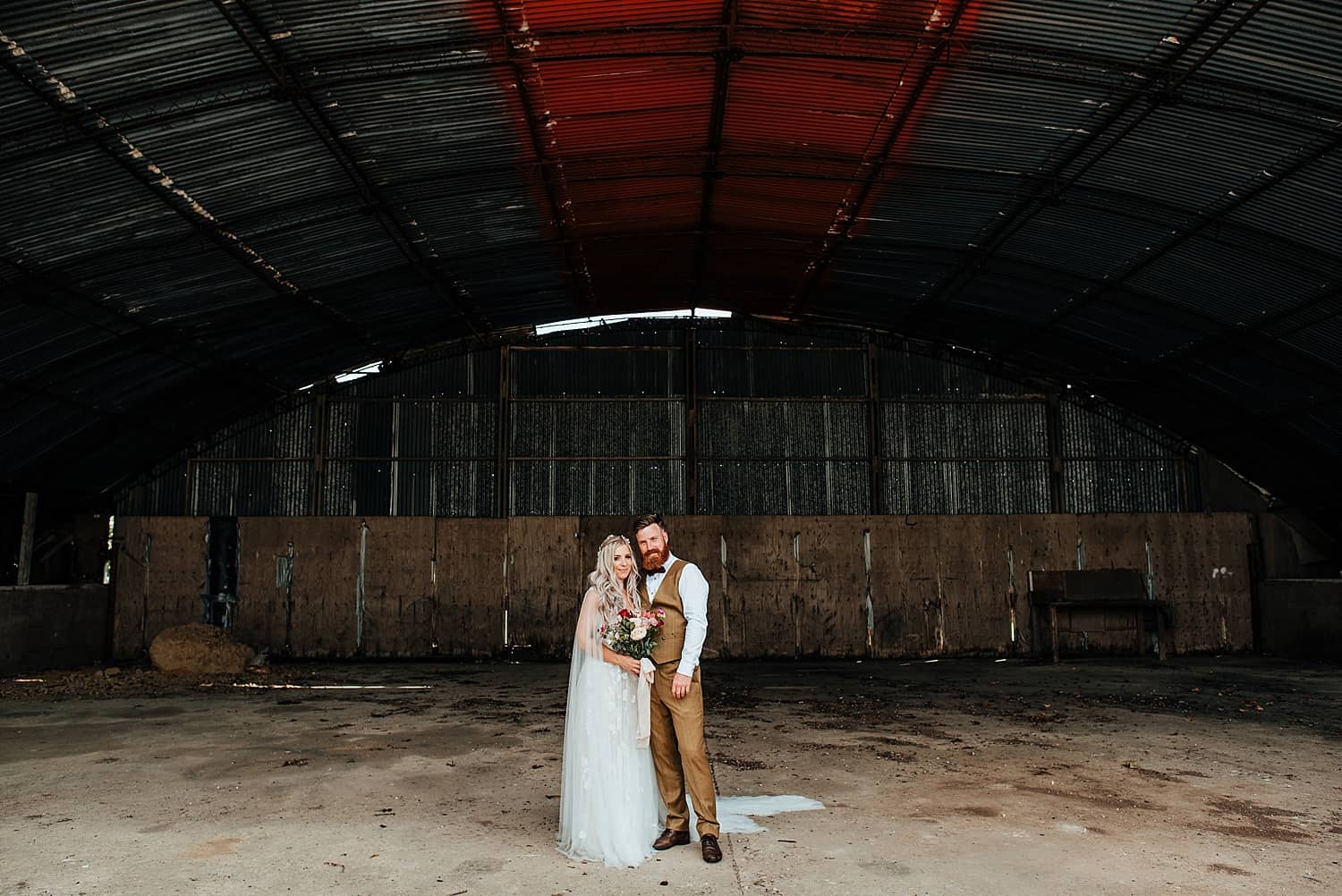 boho bride standing in an open barn with groom in brown tweed suit at clock barn